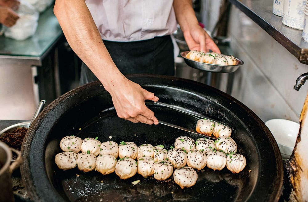 Shanghai pork bao buns. Cooked on a blackened iron fire the cook is currently lifting a group of them out of the pit using tongs and placing them into a small silver metal dish,