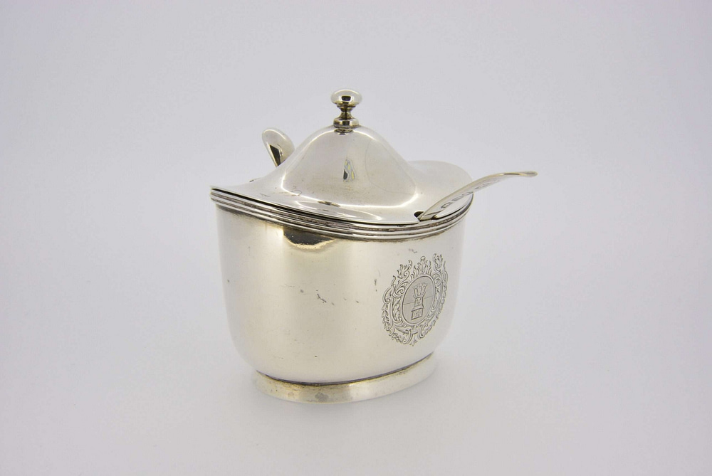 An Edward VII antique silver oval mustard pot of generous proportions, engraved with a castle crest within a foliate armorial surround with thread edging, domed hinged cover with compressed ball finial and angular handle, 8cm high, makers mark S.G for R & S Garrard and Co (Sebastian Henry Garrard) subsequently Garrard & Co, London 1906, 152gms, 4.8ozt, with original clear glass liner and a George III old English pattern mustard spoon, makers mark T.W. for Thomas Wallis, London 1808.