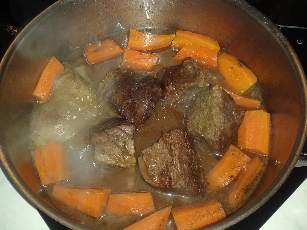 Browned chunks of beef along with sliced orange carrots cooking away in a deep, metal pan, with some onions and stock in the bottom.
