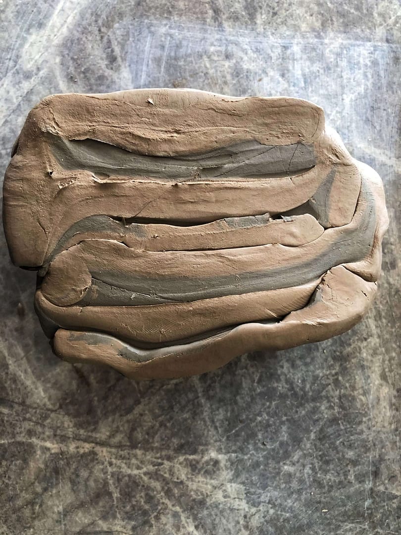 Two pieces of clay being stacked and slammed to blend them together. Both light clays but one is darker than the other. This start of the process shows the layers, like sediment in a river bed or in Arctic ice samples that as the work continues, slowly get thinner and thinner until they disappear and the clay is homogeneous.