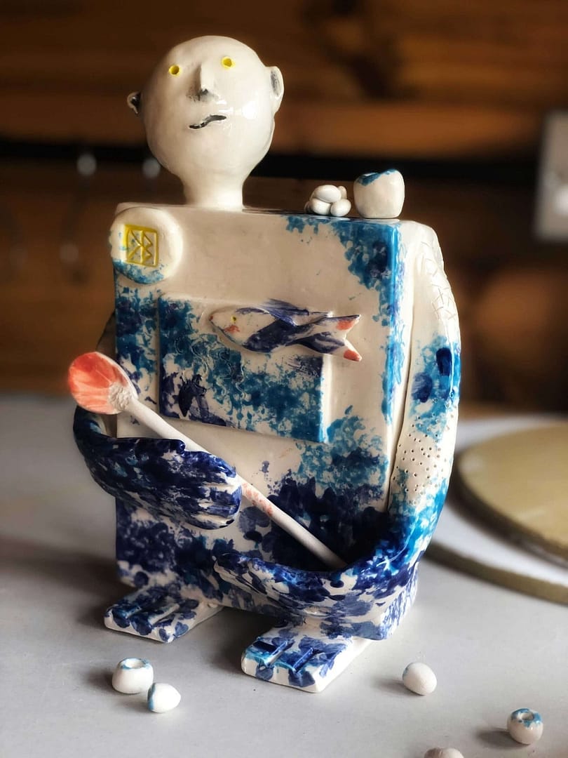 A rectangular ceramic sculpture depicting a god with long arms, blue markings, a spoon cradled in its arms, a round head set on a long neck, with strange, slabby square feet. But it's a god, it can look however it wants...