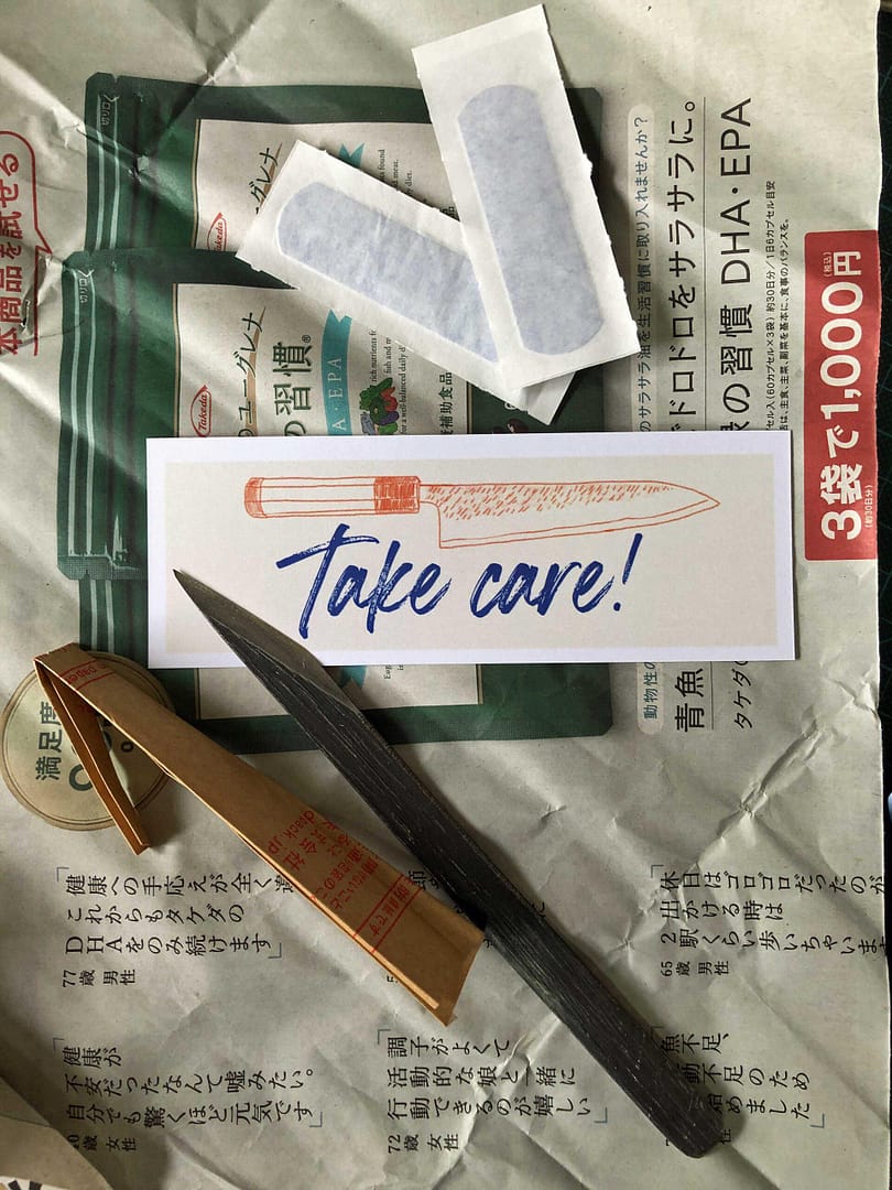 On a background of a page from a Japanese newspaper sits a small metal craft knife alongside it's brown paper safety wrapper. They also gifted two sticking plasters along with their knife care instruction card which has "Take care" on the obverse.
