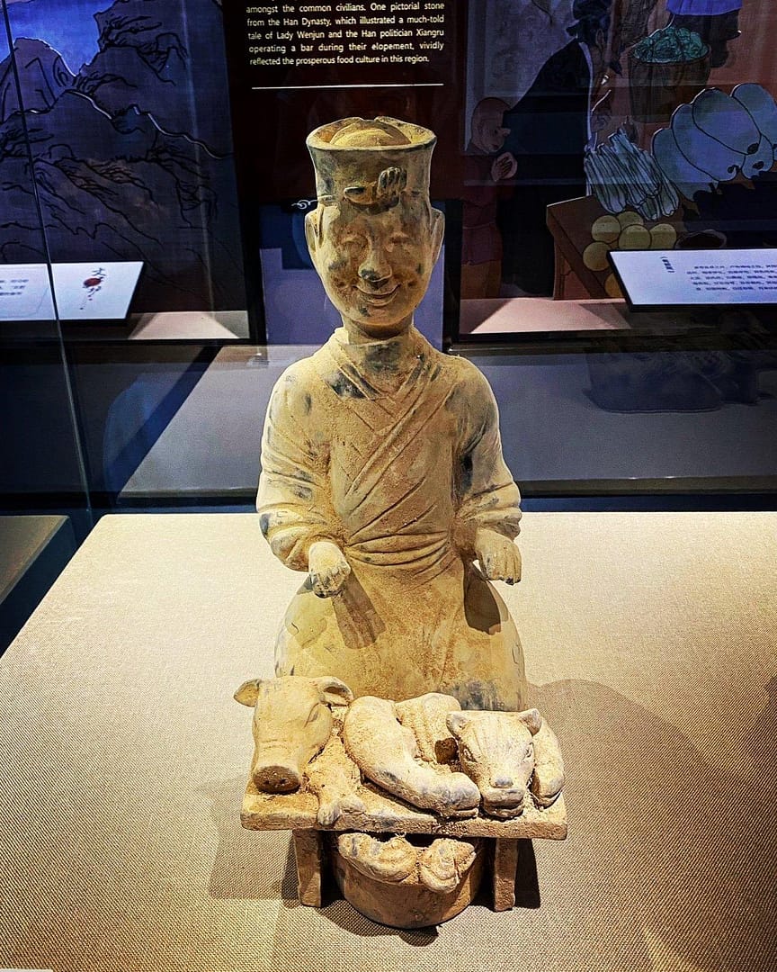 A ceramic grave offering of a chef from Han era China; it's a pale yellow/brown. The chef has a had and what I assume are traditional robes. He sits, hands out, palms upwards. In front of him is a low table with a number of animal heads and a basket underneath the table that looks to contain loaves (?)