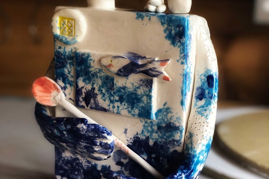 A rectangular ceramic sculpture depicting a god with long arms, blue markings, a spoon caredeled in its arms, a round head set on a long neck, with strange, slabby square feet. But it's a god, it can look however it wants...