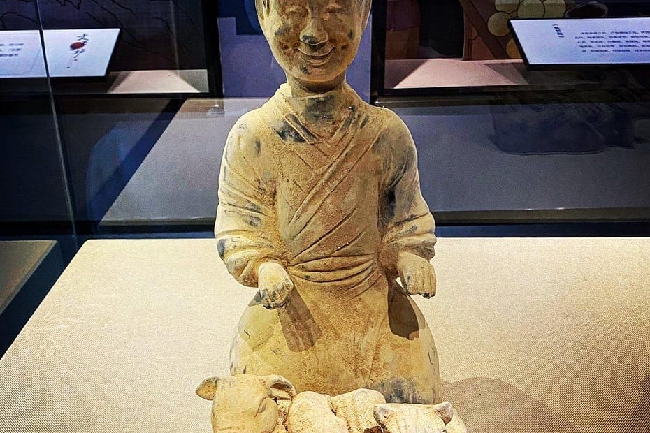 A ceramic grave offering of a chef from Han era China; it's a pale yellow/brown. The chef has a had and what I assume are traditional robes. He sits, hands out, palms upwards. In front of him is a low table with a number of animal heads and a basket underneath the table that looks to contain loaves (?)