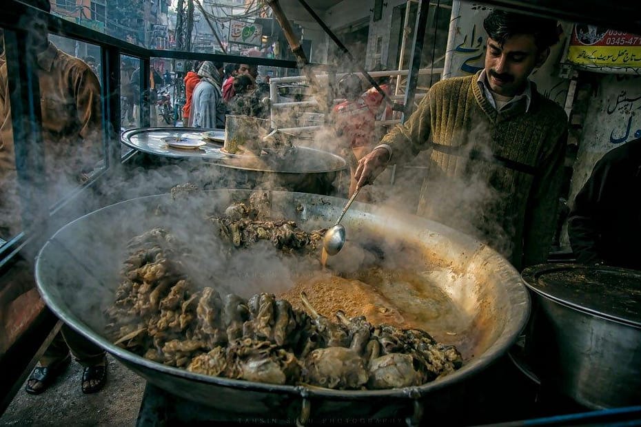 A cook in Lahore, Pakistan dressed in a brown, V-neck pullover is stirring a large flat pan of Siri Payey (Head and feet goat curry) with a large spoon which is a very popular cuisine of Pakistan' traditional food for breakfast. Steam rises off the cooking meat & in the background, through the misted windows, you can see large crowds of people moving back & forth through what is obviously a partially covered market area.
