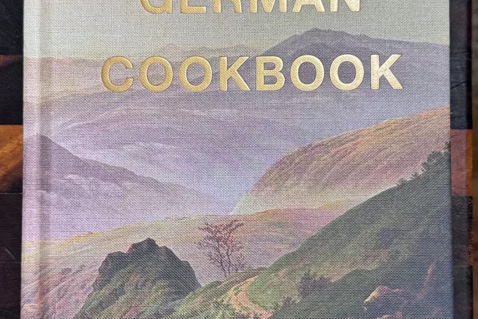 Front cover of The German Cook book, showing a blue, misty mountain side and valley with other hills rising and falling in the distance. A rocky track winds down the side with small sparse trees on each side. There are goats wandering around on the left hand side.