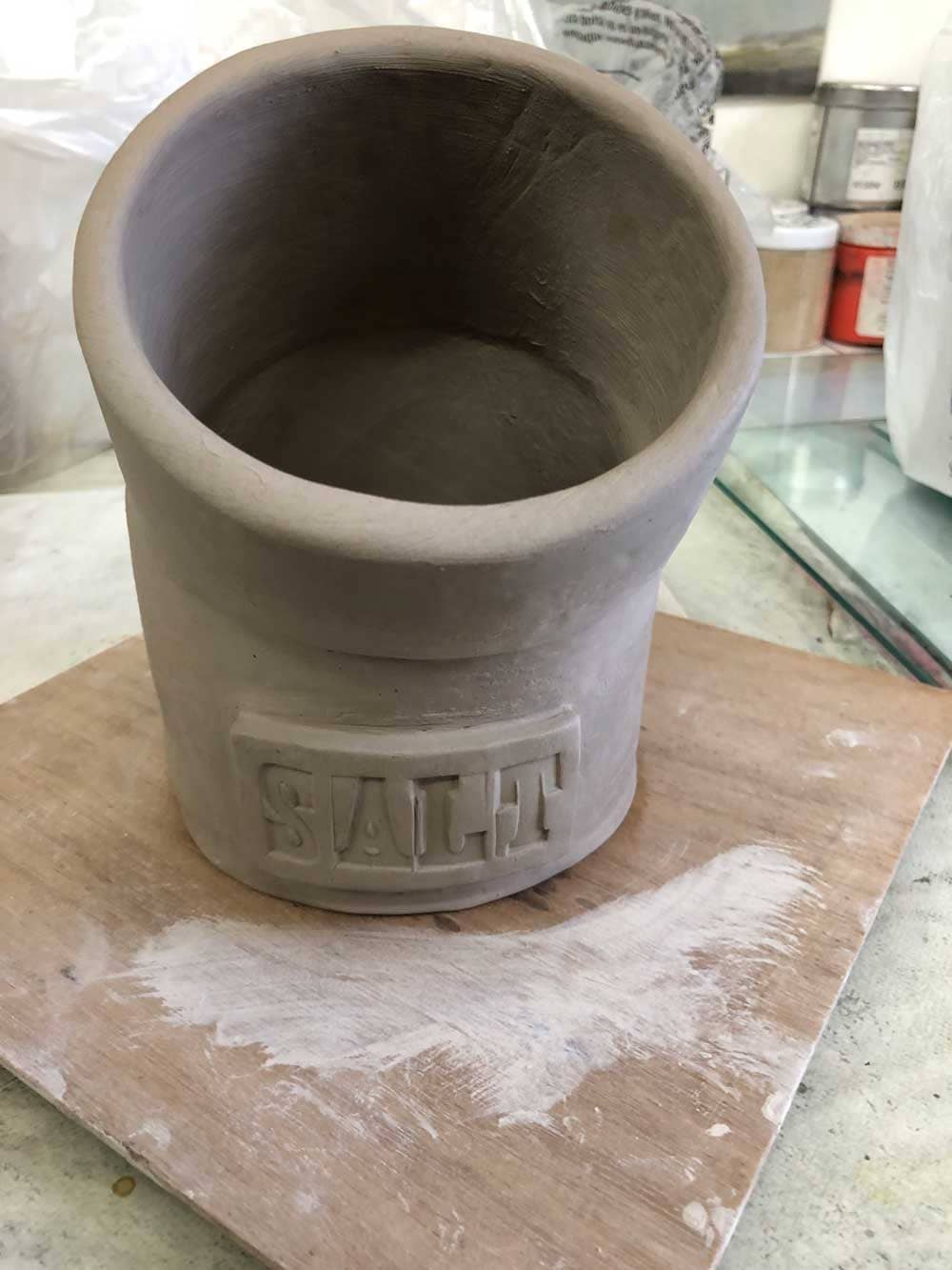 A salt-pig, currently unfired, so grey/brown in colour, with a little name-plate reading "SALT" at the base. It's made up of one cylinders, cut at an angle of about 30 degrees into two, one is rotated and then they're both re-joined.
