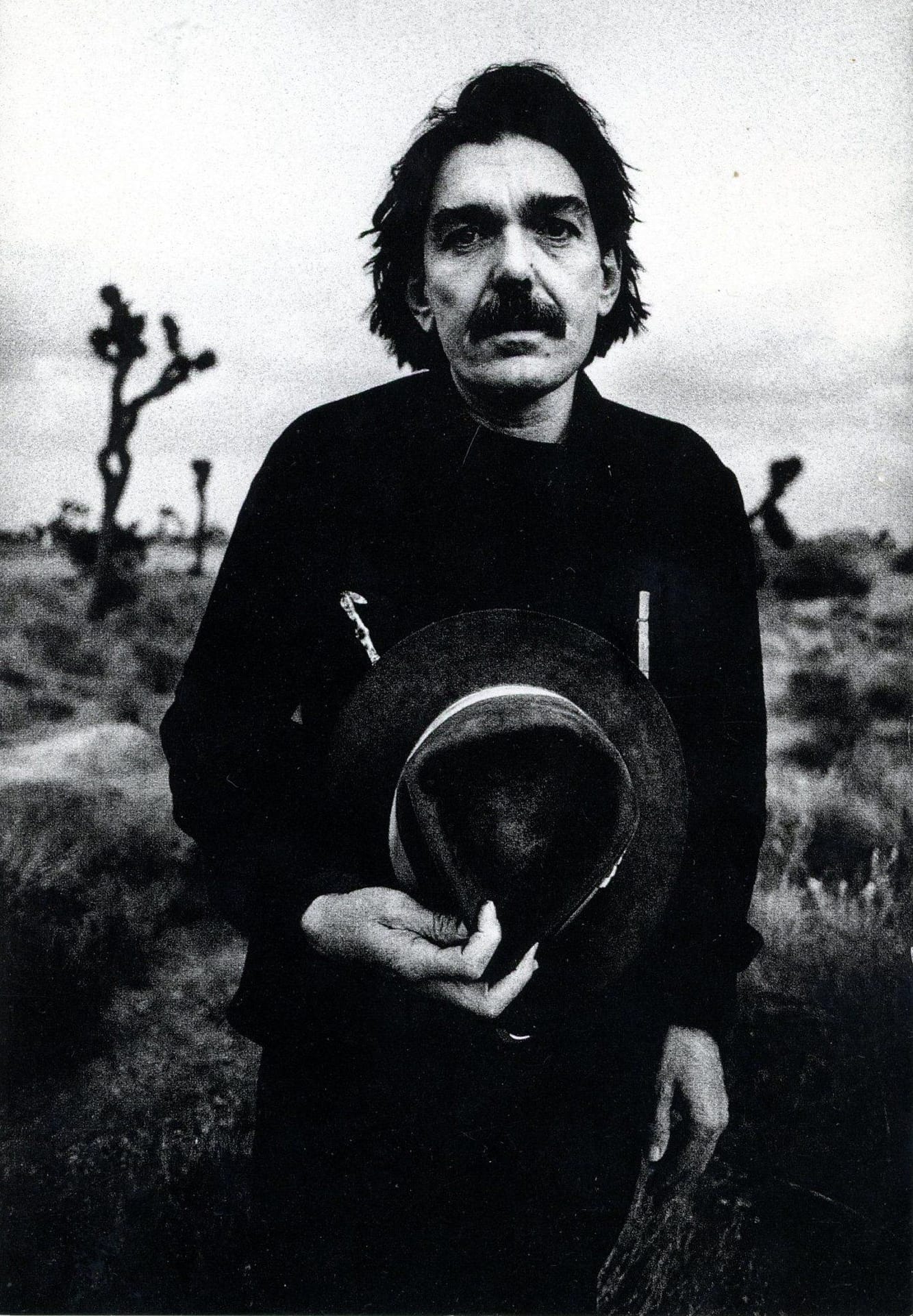 A classic B&W photo of Don van Vliet (Captain Beefheart) by photographer Anton Corbijn taken in the Mojave desert. He's holding one of his wide-brimmed hats against his chest, eyes sad, black hait, large dark moustache and bushy eyebrows, cacti thrusting up in the background with a boulder just behind him.