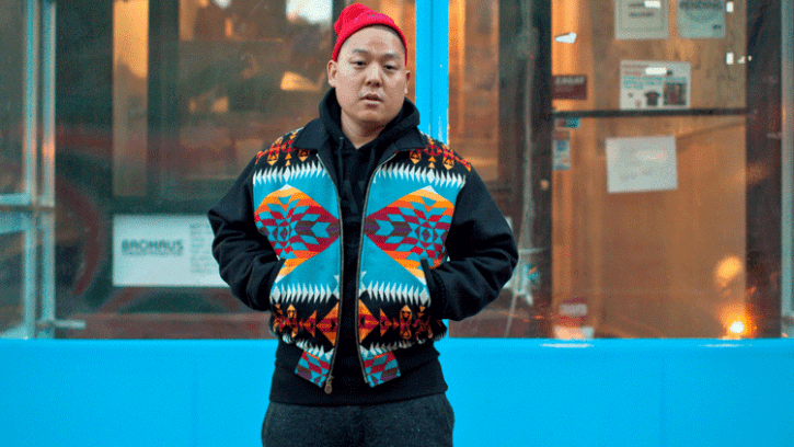 Chef Eddie Huang wearing a multi-coloured woollen jacket, standing in front of an unidentified shop front, with his hands thrust into the pockets and with a red beanie on this head.