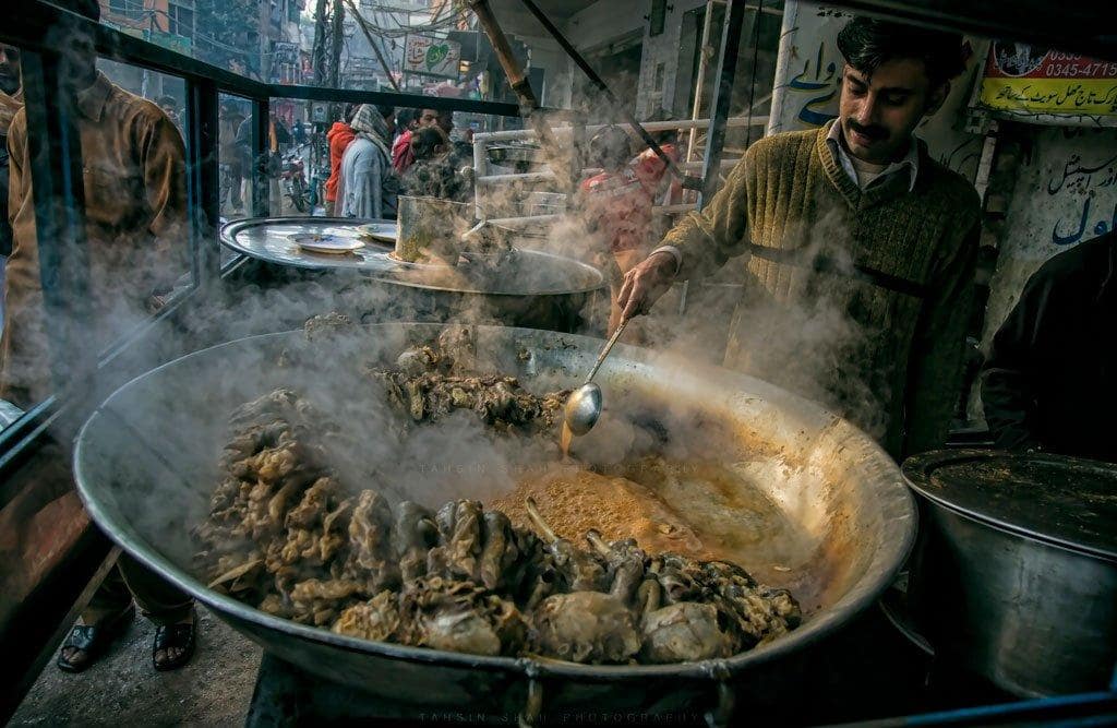 A cook in Lahore, Pakistan dressed in a brown, V-neck pullover is stirring a large flat pan of Siri Payey (Head and feet goat curry) with a large spoon which is a very popular cuisine of Pakistan' traditional food for breakfast. Steam rises off the cooking meat & in the background, through the misted windows, you can see large crowds of people moving back & forth through what is obviously a partially covered market area.