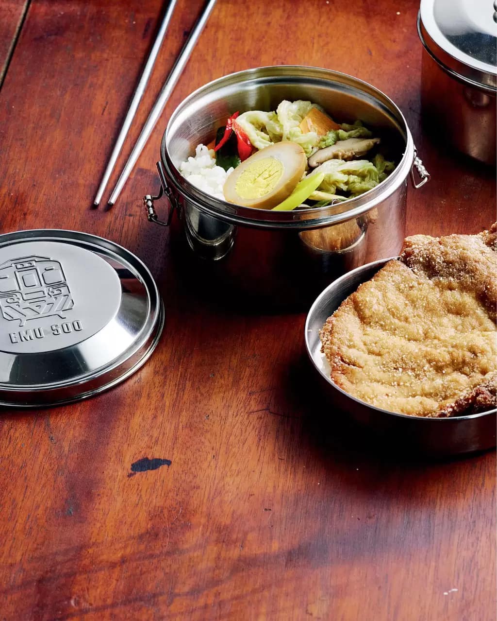 A fried pork chop in a stainless steel bento box, with rice and pickles and salad in the other half, and a pair of metal chopsticks.