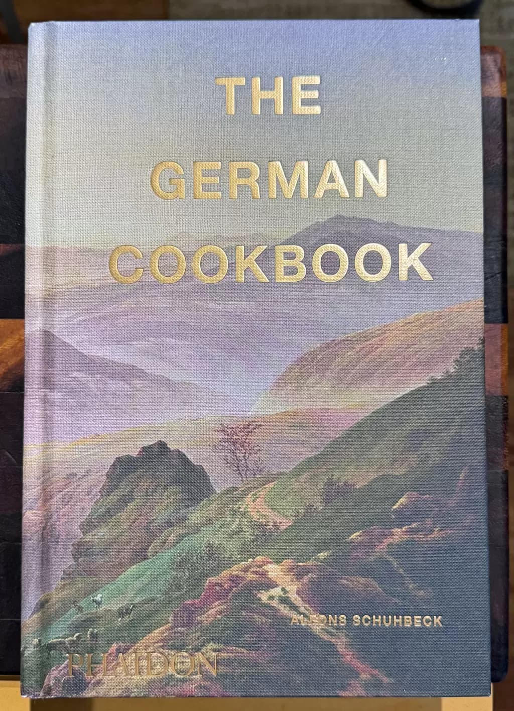 Front cover of The German Cook book, shoing a misty muntain side and valley with other hills rising and falling in the distance. A rocky track winds down the side with small sparse trees on each side.