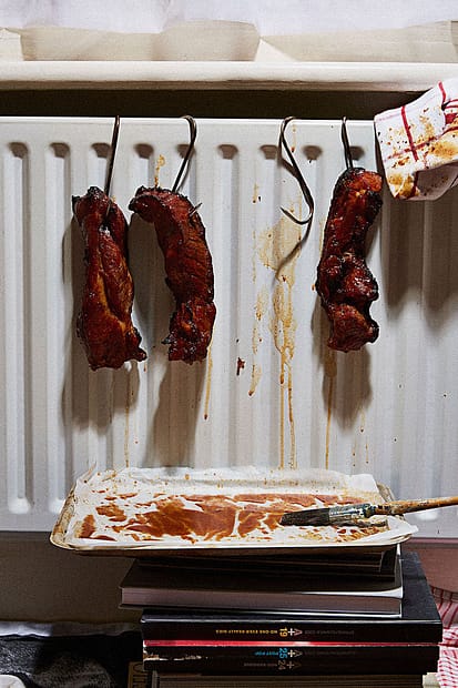 Dark red, pork belly chunks being hung over a white radiator on butchers hooks, with sauce dripping down off it onto a tray stationed underneath, which is lifted off the ground on a stack of magazines. A dirty kitchen towel is draped alongside the meats.