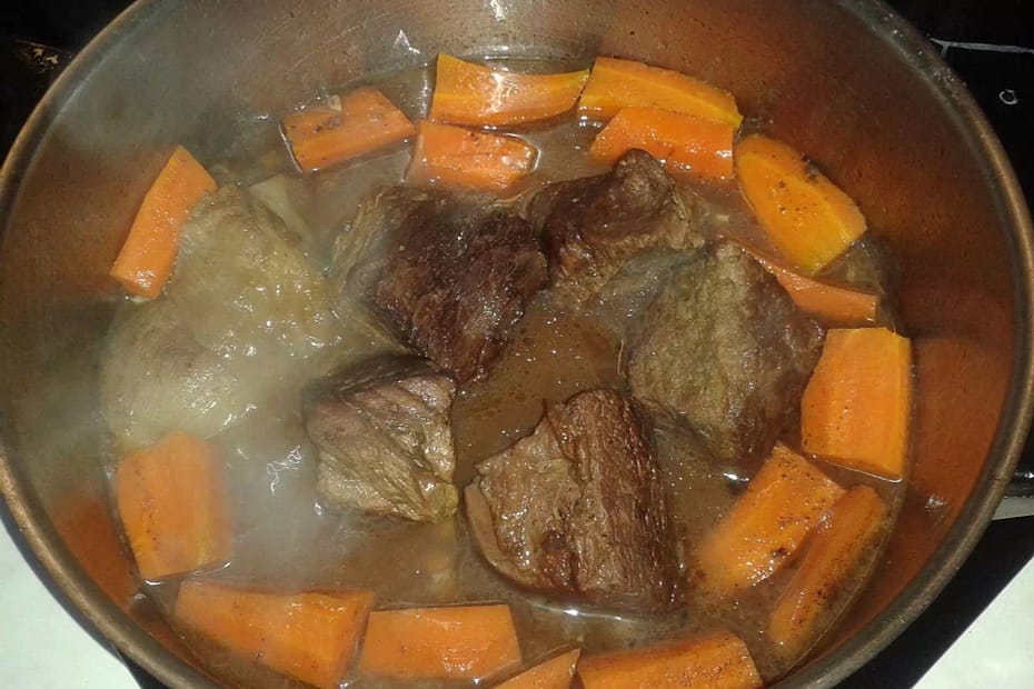 Browned chunks of beef along with sliced orange carrots cooking away in a deep, metal pan, with some onions and stock in the bottom.
