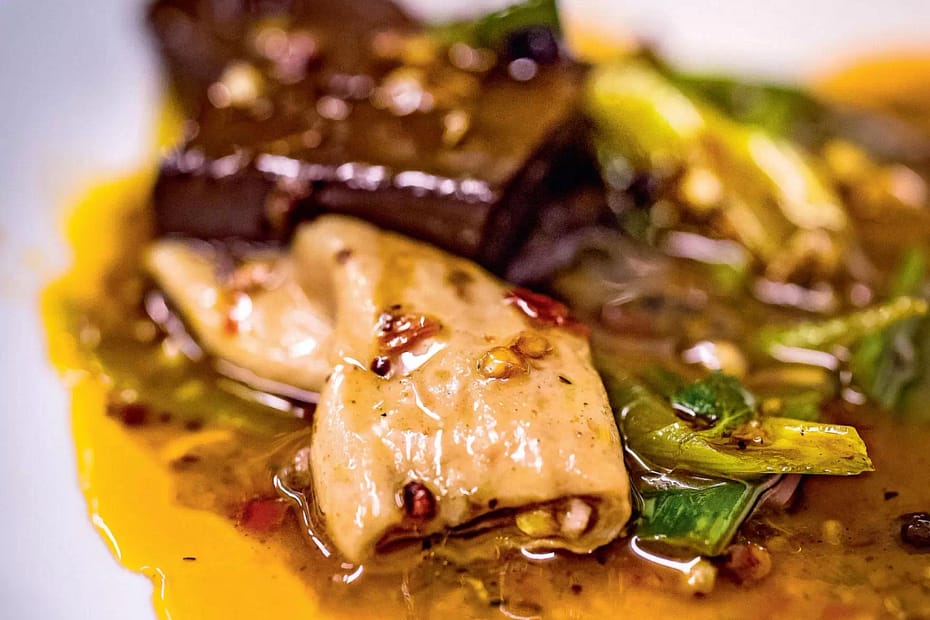 yellow and red oily sauce surrounds a lightly cooked and browned intestine with green mustard leaves all on a white plate.
