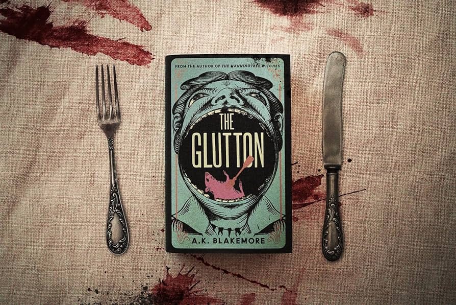The book, "The Glutton" set against a blood-stained sacking table cloth, flanked by antique silver looking knife & fork. The book shows a cartoon head, almost completely overshadowed by the teeth of the man filling the centre of the cover, inside of which resides a, dead, red rat.