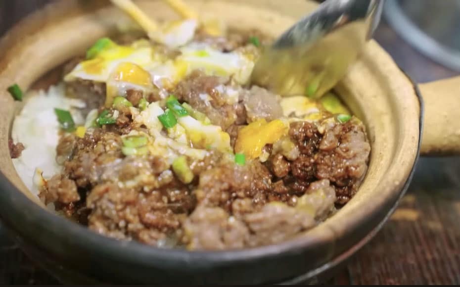 A bowl of rice with beef topping, stirred together in a ceramic rice-pot. A fried egg has been stirred into the mix and a spoon sits on the right side, ready to eat this delight.