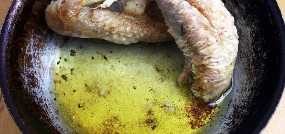 Part of a chicken being cooked off, with the golden fat already rendering out into the scratched bottom of an old frying pan.