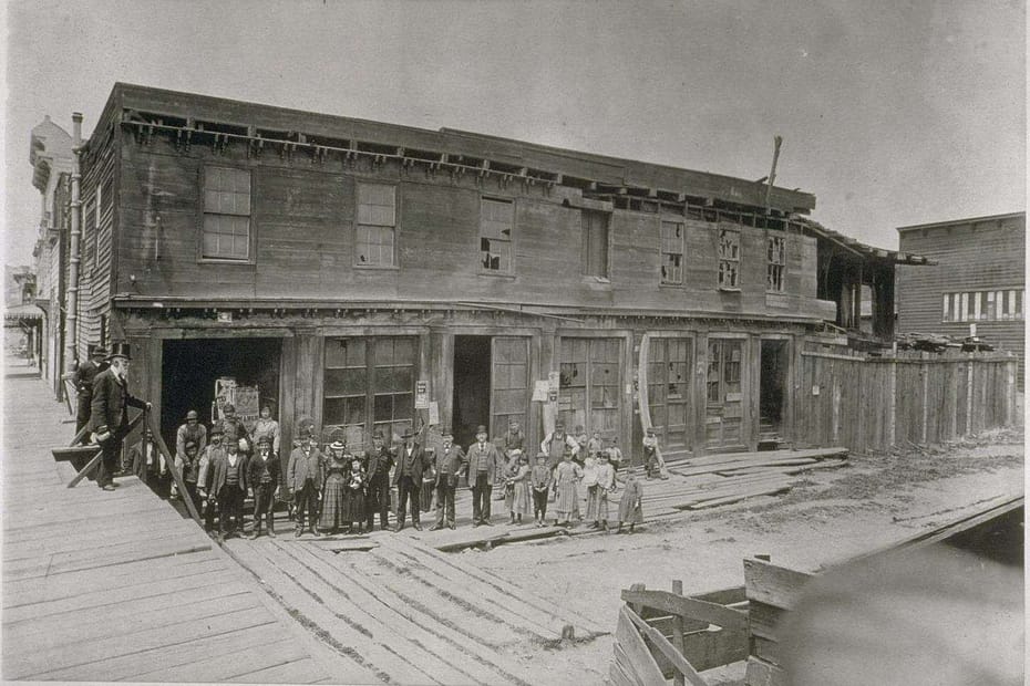 An old, large, wooden built barn-like structure, unpainted, dirty, almost derelict with a group of old-timers standing in the front.